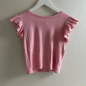 Rosa ribbad topp med volangärmar - Söt rosa topp i ribbad bomull med volangdetaljer på ärmarna. Det är en liten gul fläck på magen som borde gå bort vid tvätt. Perfekt för vår och sommar när du vill ha något lätt och luftigt🩷🩷