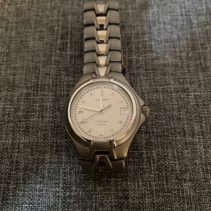 Tissot herrklocka i titan - Stilren herrklocka från Tissot i titan med silverfärgad urtavla och datumvisning. Armbandet är robust och matchar boetten i samma material. Klockan är vattentät upp till 200 meter och har safirglas.