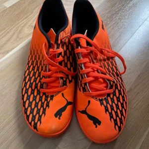 Orangea fotbollsskor från Puma - Säljer ett par orangea fotbollsskor från Puma med svart mönster och logga på sidan och tån. Skorna har snörning och är designade för att ge bra grepp på planen. Perfekta för dig som vill sticka ut på matchen!