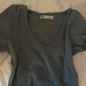 Grön ribbad topp från Pull&Bear - Säljer en grå/grön ribbad topp från Pull&Bear med v-ringning och korta puffärmar. Perfekt till jeans eller kjol för en enkel och snygg look.