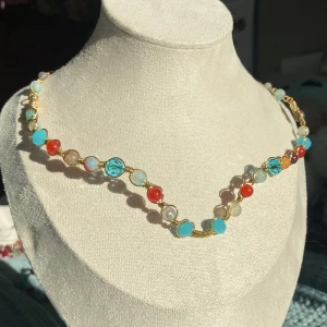 Färgglatt guldhalsband med pärlor - Unikt halsband med runda pärlor i blått, rött, vitt och beige, trädda på en tunn guldfärgad kedja. Perfekt för att ge din outfit en färgklick och personlig touch. Smycket är flexibelt och går att forma efter sin egen hals. Finns i silver också. 