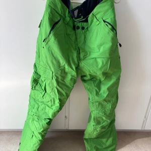 Gröna täckbyxor från Stuf - Säljer ett par gröna täckbyxor från Stuf i storlek L. Byxorna har svarta detaljer vid fickor och midja, dragkedjor och knappar framtill samt flera praktiska fickor. Perfekta för vinterns alla äventyr i snön.