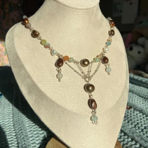 Halsband med bruna pärlor och färgade stenar - Unikt halsband med stora bruna pärlor och små runda stenar i blått, grönt och orange. Silverfärgad kedja med dekorativa hängande detaljer framtill. Perfekt statement-piece för dig som gillar färg och personliga smycken.