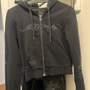 Svart hoodiejacka från Ed Hardy - Svart hoodiejacka från Ed Hardy med dragkedja framtill och tryckt logga på bröstet. Stora fickor fram och coolt tatueringsinspirerat motiv på ryggen. Justerbar huva med snören och ribbade muddar.