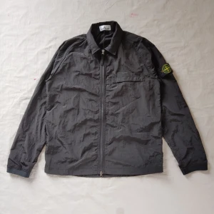 Grå overshirt från Stone Island - Säljer en grå overshirt från Stone Island med klassisk krage och dragkedja framtill. Jackan har lång ärm, bröstficka och den ikoniska loggan på vänster ärm. Materialet är lätt och syntetiskt, perfekt för lager-på-lager.