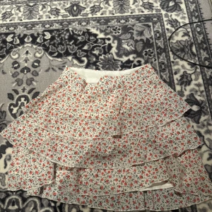 Blommig volangkjol med små röda blommor - Supersöt kort kjol med volanger och blommigt mönster i rött och grönt på vit botten. Perfekt för dig som gillar romantisk och lekfull stil. Kjolen har flera lager volanger som ger extra volym och rörelse.