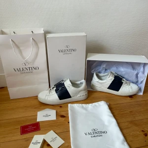 Valentino Garavani Open mörkblå  - Säljer ett par vita Valentino Open sneakers med bred mörkblå  läderdetalj över mitten. Snygg och stilren design som passar till mycket. Skorna är i skinn och kommer med originalpåse, box och äktehestkort.                                                     Storlek 44 men passar 43.                                   9/10 skick!! 
