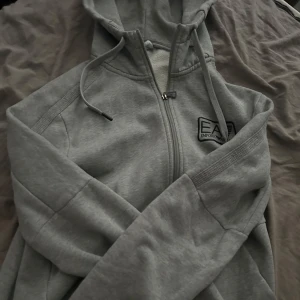 Grå hoodie från EA7 Emporio Armani - Säljer en grå hoodie från EA7 Emporio Armani med dragkedja och huva. Jackan har lång ärm, ribbade muddar och en stilren logga på bröstet. Perfekt för en avslappnad och sportig look. Nypris 1899 jag säljer nu den för runt 800kr