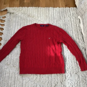 Röd kabelstickad tröja från GANT.  - Säljer en röd kabelstickad tröja från GANT med rund halsringning. Tröjan har långa ärmar och diskret logga broderad på bröstet. Klassisk och stilren modell som passar perfekt till kyligare dagar.