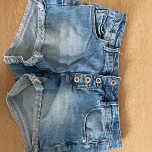 Blå jeansshorts med knappar - Säljer ett par klassiska blå jeansshorts med fransiga benslut och synliga knappar framtill. Shortsen har fem fickor och en snygg tvättad look. Perfekta för sommaren och enkla att matcha med allt. Säljer för att det tu är inte passar. Pris kan diskuteras.