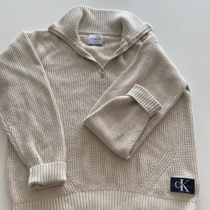 Beige stickad tröja med dragkedja från Calvin Klein Jeans - Säljer en beige stickad tröja från Calvin Klein Jeans med halv dragkedja och hög krage. Tröjan har ribbade muddar och en svart CK-logga framtill. Perfekt för lager-på-lager och stilren look. 