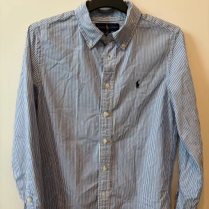 Blåvit randig skjorta från Ralph Lauren - Klassisk blåvit randig skjorta från Ralph Lauren med button-down krage och broderad logga på bröstet. Skjortan har långa ärmar och knäppning framtill. Perfekt för en stilren look.