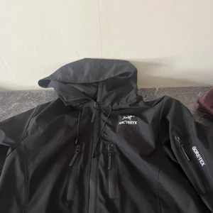 Svart skaljacka från Arc'teryx med huva - Säljer en svart skaljacka från Arc'teryx i storlek M. Jackan har huva, dragkedja framtill och GORE-TEX-material som gör den perfekt för blåsiga och regniga dagar. Snygg och funktionell med logga på bröstet och ärmficka.