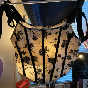 Vit och svart blommig korsett-topp med knytband - Säljer en snygg korsett-topp i vitt med svarta blommor och svarta vertikala ränder. Toppen har knytband på axlarna och en croppad passform. Perfekt för att lyfta vilken outfit som helst! Aldrig använd 