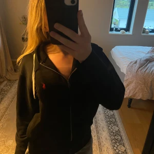 Svart hoodie från Polo Ralph Lauren - Detta är en svart hoodie med dragkedja från Polo Ralph Lauren + luva. Det är en väldigt fin tröja som tyvärr bara legat i garderoben, jag hoppas någon annan har användning av den🫶🏻💕köpt för 1100kr