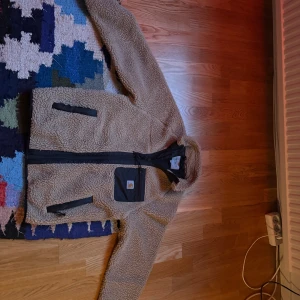 Beige flis jacka från Carhartt - Mysig beige flis jacka från Carhartt med svarta detaljer och dragkedjor. Jackan har en stor svart bröstficka med Carhartt-logga och två sidofickor med dragkedja. Perfekt för kyliga dagar och riktigt skön att ha på sig.