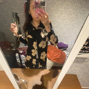 Svart blommig skjortklänning från Wocci - Säljer en svart skjortklänning från Wocci med stora beige blommor och lila detaljer. Klänningen har lång ärm, krage och knäppning framtill. Perfekt för dig som gillar statement-mönster och avslappnad stil.