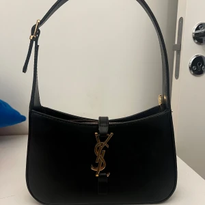 Svart axelväska från Saint Laurent - Snygg svart axelväska från Saint Laurent med guldiga detaljer och klassisk YSL-logga framtill. Väskan har justerbart axelband med spänne och dragkedja upptill. Perfekt för att lyfta vilken outfit som helst.