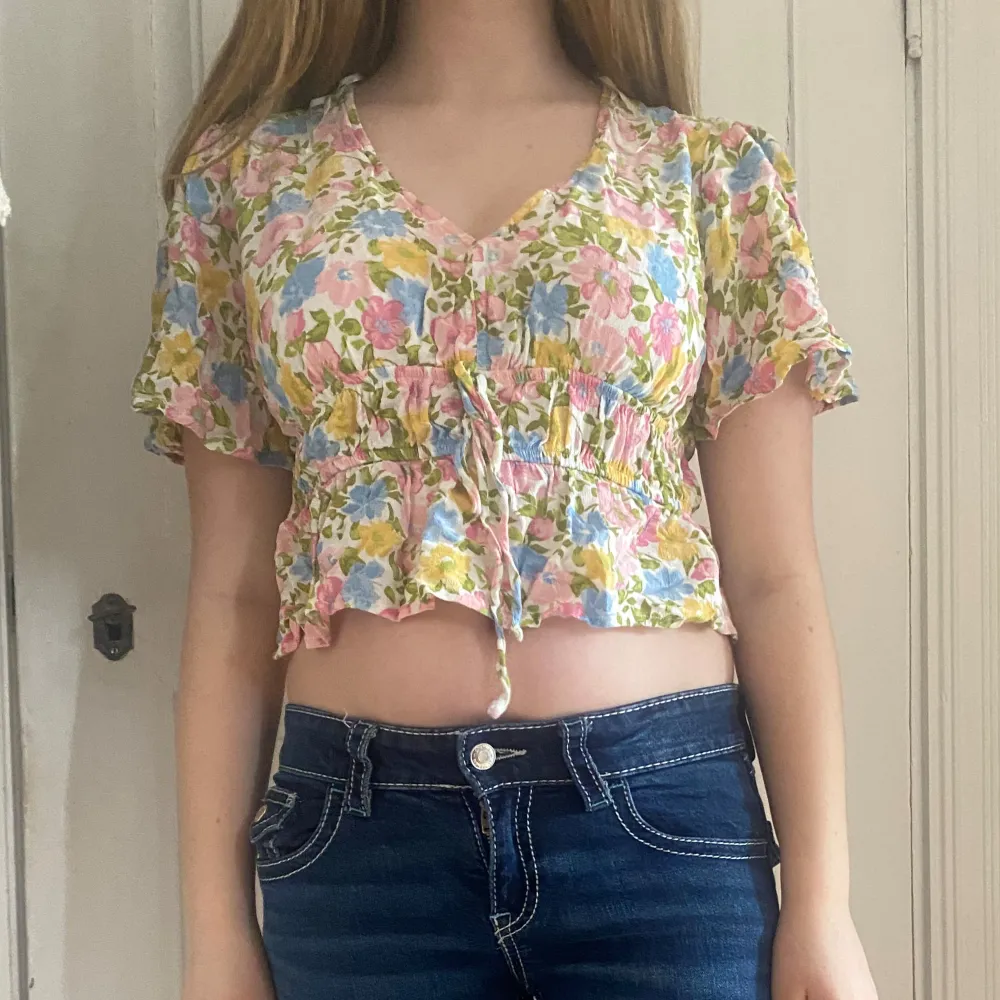 Söt blommig blus i pastellfärger med korta ärmar och v-ringning. Toppen är croppad och har knytning framtill samt volangdetaljer vid midjan och ärmslut. Perfekt för vår och sommar!. Yläosat.