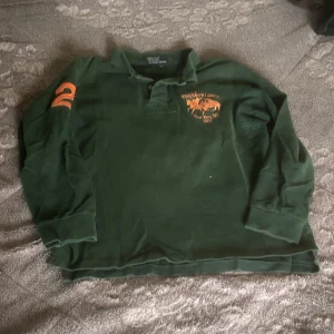 Grön pikétröja från Polo Ralph Lauren - Mörkgrön långärmad pikétröja från Polo Ralph Lauren med orange broderad logga på bröstet och siffran 2 på ärmen. Klassisk krage och knäppning framtill. Custom fit-modell.