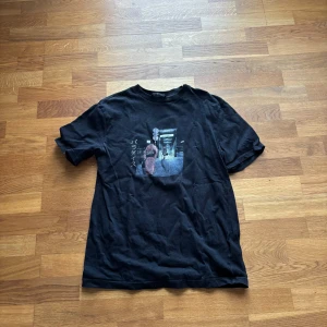 Svart t-shirt med japanskt tryck - Svart t-shirt med ett tryck framtill som föreställer en person i traditionell japansk klädsel och japanska tecken. T-shirten har rund hals och korta ärmar. Perfekt för dig som gillar streetwear med asiatisk touch.