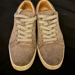 Gråa sneakers från Christian Louboutin - Säljer ett par gråa sneakers från Christian Louboutin med klassisk röd sula. Skorna har snörning och är tillverkade i mocka med vita kanter runt sulan. Perfekta för dig som gillar stilrena och exklusiva sneakers.priset är ej hugget i sten och kan få till baka sitt glans med lite putsning