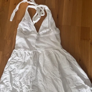 Vit  halterneckklänning - Säljer en vit kortklänning med halterneck och v-ringning. Klänningen har ett smockat midjeparti och  broderade detaljer. Perfekt för varma dagar och midsommar🥰