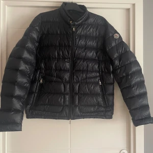 Svart pufferjacka från Moncler - Säljer en svart pufferjacka från Moncler med hög krage och dragkedja framtill. Jackan har två fickor med dragkedja och Moncler-logga på ärmen. Klassisk quiltad design och lätt glansigt material.
