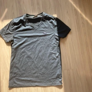 Grå t-shirt med svarta ärmar från Lacoste - Säljer en grå t-shirt från Lacoste med svarta ärmar och v-ringad hals. Klassisk krokodillogga broderad på bröstet. Tillverkad i 100% bomull och har en normal passform. Perfekt för en stilren och avslappnad look.