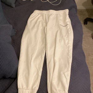 Vita mjukisbyxor från Lacoste - Säljer ett par vita mjukisbyxor från Lacoste med resår i midjan och vid bensluten. Byxorna har en diskret Lacoste-logga på ena benet och är tillverkade i mjuk bomull, perfekta för en avslappnad stil.