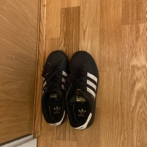 Svarta Adidas Superstar sneakers - Klassiska svarta Adidas Superstar sneakers med vita ränder och snörning. Skorna har rund tå och platt sula, samt den ikoniska gummitåhättan. Perfekta för en sportig och avslappnad stil. pris kan diskuteras 
