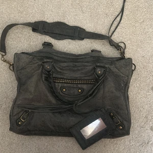  Balenciaga city bag - Cool mörkgrå axelväska från Balenciaga med klassisk design och detaljer i guld. Tillhörande spegel och flera fack för smart förvaring. Handtagen är i lite dåligt skick och väskan har en liten hål(syns inte I bruk), titta 3 bilden. Har äktighäts bevisning. Tar emot prisförslag.
