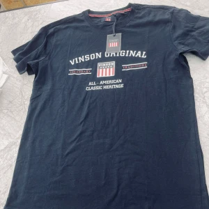 Mörkblå t-shirt från Vinson - Snygg mörkblå t-shirt från Vinson Original med vit text och tryck framtill. Klassisk rund halsringning och korta ärmar. Perfekt för en avslappnad stil.