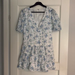 Blommig kortklänning med volang - Superfin vit kortklänning med blått blommönster och volanger. Klänningen har v-ringning och korta puffärmar, totallängd 75cm, ej använd 