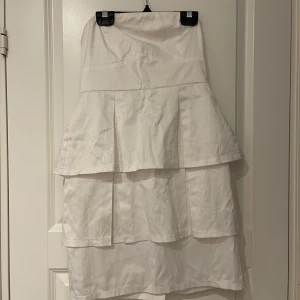 Vit axelbandslös klänning med volanger - Säljer en vit axelbandslös klänning från Miss Etam med två lager volanger och dragkedja bak. Klänningen har en elegant och stilren design, perfekt för sommarens tillfällen. Materialet känns mjukt och har en lätt glans.