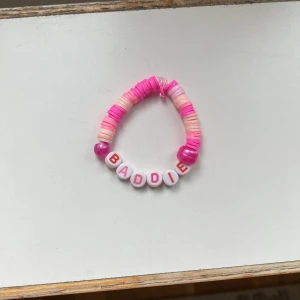 Armband med bokstavspärlor 'BADDIE' i rosa och vitt gjort själv! ❤️ - Säljer ett färgglatt armband med platta pärlor i olika nyanser av rosa och vitt. Armbandet har vita bokstavspärlor som bildar ordet 'BADDIE' och är elastiskt för enkel passform. Perfekt accessoar för att addera lite attityd till din stil! Den ser liten ut men det är elastisk tråd så den stretchar ut för din handled skulle nog rekommendera i åldern 6-12