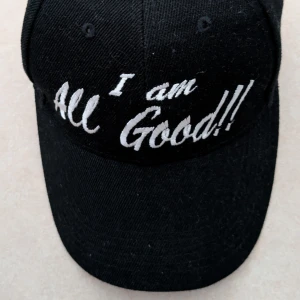 Svart keps med vit brodyr - Svart keps med böjd skärm och justerbar rem bak. Framsidan har vit brodyr med texten  I am All Good!!i en cool, handskriven stil. Perfekt accessoar för en avslappnad look. Kepsen har aldrig använts 