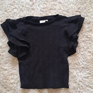 Svart volangtopp från Gina Tricot - Säljer en svart topp från Gina Tricot med snygga volangdetaljer på ärmarna. Toppen har en ribbad struktur och korta ärmar med extra volym. Perfekt för dig som vill ha något stilrent men ändå med en twist.