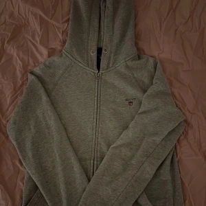Grå hoodie med dragkedja från GANT - Grå hoodie från GANT med dragkedja framtill och liten broderad logga på bröstet. Mjuk bomullskänsla och huva med snörning. 
