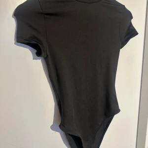 Svart kortärmad bodysuit - En enkel och stilren svart bodysuit med korta ärmar och rund halsringning. Perfekt att styla med jeans eller kjol för en clean look. Gjord i mjukt och stretchigt material för en bekväm passform.