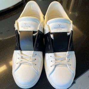Valentino Garavani sneakers - Snygga vita Valentino Garavani sneakers med en bred svart rem över snörningen. Skorna har en stilren design med subtila detaljer och är perfekta för en trendig look. Pris kan diskuteras om du gör en snabb affär