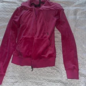Rosa velour hoodie från Juicy Couture - Juicy Couture hoodie i färgen raspberry, velour med dragkedja och huva. Klassisk broderad JC-logga på bröstet och fickor framtill. Mjuk och skön med ribbade muddar vid ärmslut och nederkant. Perfekt för en avslappnad och trendig look.                 Har några små bränn märken, ej hål. Hoodien är xs och byxorna xxs   