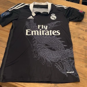 Real Madrid svart fotbollströja med drakmotiv - Säljer en svart Real Madrid fotbollströja från Adidas med vitt drakmotiv på framsidan. Tröjan har vita ränder på axlarna, vit krage och tryckta loggor samt Champions League-märken på ärmarna. Fly Emirates sponsortryck på bröstet.