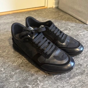  Valentino rockrunner - Säljer ett par svarta Valentino Garavani rockrunner sneakers med diskret camouflage-mönster. Skorna har snörning, rund tå och platt sula. Materialmix av mocka och skinn ger en lyxig känsla. Perfekta för dig som vill ha stilrena och exklusiva sneakers. box ingår!