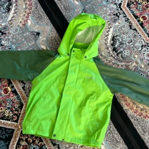 Grön regnjacka med huva från Exani - Säljer en grön regnjacka från Exani med huva och reflexdetaljer. Jackan har mörkgröna ärmar, dragkedja och tryckknappar framtill samt en reflexstjärna på ryggen. Perfekt för regniga dagar. För barn.