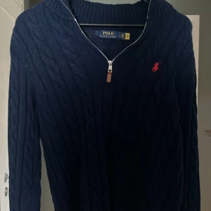 Mörkblå stickad tröja med dragkedja från Polo Ralph Lauren - Säljer en mörkblå stickad tröja från Polo Ralph Lauren med halv dragkedja och klassiskt kabelstickat mönster. Tröjan har lång ärm och röd broderad logga på bröstet. Perfekt för en stilren och avslappnad look.