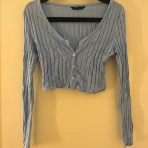 Ljusblå croppad kofta från SHEIN - Säljer en ljusblå croppad kofta från SHEIN med ribbad struktur och v-ringning. Tröjan har långa ärmar och knäppning framtill med små vita genomskinliga knappar. Perfekt för lager-på-lager och en chill look.