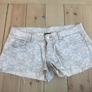 Blommiga jeansshorts med fransar - Säljer ett par ljusa jeansshorts med blommigt mönster i vitt och blått. Shortsen har råa fransade kanter, fem fickor och knappgylf. Perfekta för sommaren och har en snygg, avslappnad look.