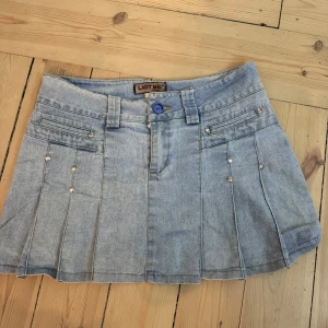 Ljusblå jeanskjol från Lady Mei - Säljer en ljusblå jeanskjol från Lady Mei med kort längd och dekorativa nitar framtill. Kjolen har fickor både fram och bak och en blå knapp i midjan. Mått finns bland bilderna