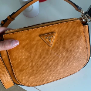 Orange axelväska från Guess - Snygg orange axelväska från Guess med strukturerad yta och klassisk trekantslogga framtill. Väskan har justerbar axelrem, dragkedja upptill och extra fack med dragkedja på baksidan. Insidan är ljus med Guess-tryck och flera fack för småsaker.